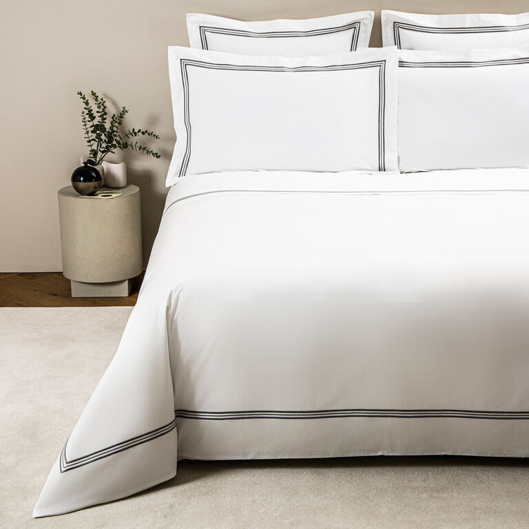 A neatly made bed features the Frette Triplo Bourdon | Duvet Cover in white cotton poplin with thin black three-line embroidery, topped with two stacked pillows and paired with a round nightstand holding a vase with greenery.