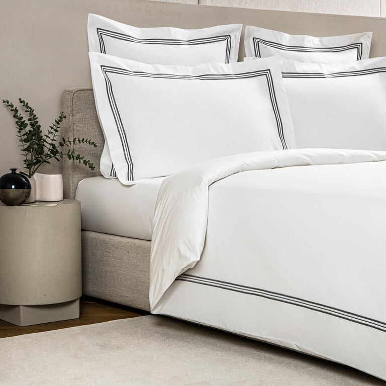 A neatly made bed features the Frette Triplo Bourdon Duvet Cover in white cotton poplin with black double-line borders, two stacked pillows, and a bedside table with a black vase and green leaves.