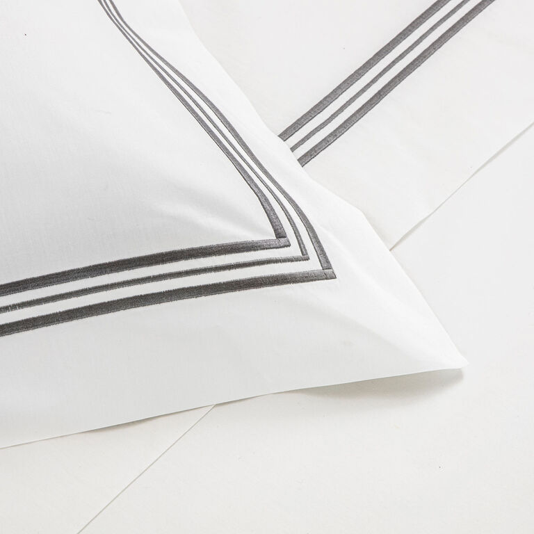 A close-up of the Frette Triplo Bourdon Pillow Sham in white cotton poplin with a double grey border and subtle sheen, elegantly displayed on a white bed.