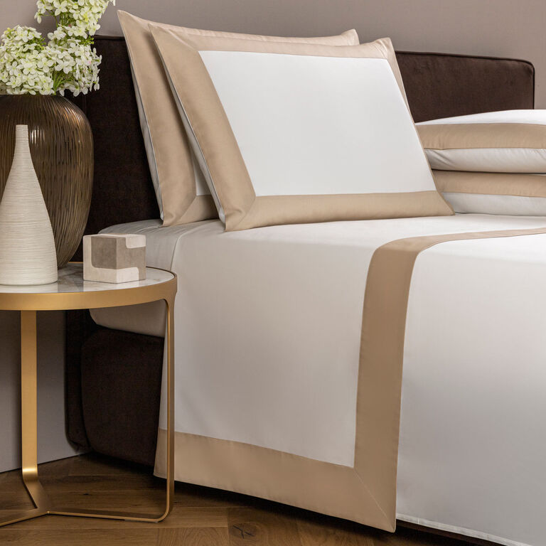 A neatly made bed with the Frette Bold | Sheet Set in white and beige borders adds modern style to the bedroom, complemented by a round gold side table with a vase of white flowers and decorative accents.