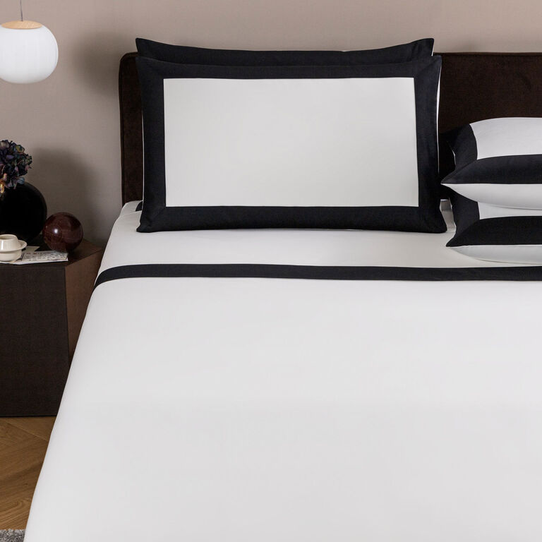 A bed dressed in Frette’s Bold | Sheet Set—crisp white sheets and pillowcases with striking black borders—creates a modern vibe, complemented by a bedside table holding a cup, saucer, and a small vase of flowers.
