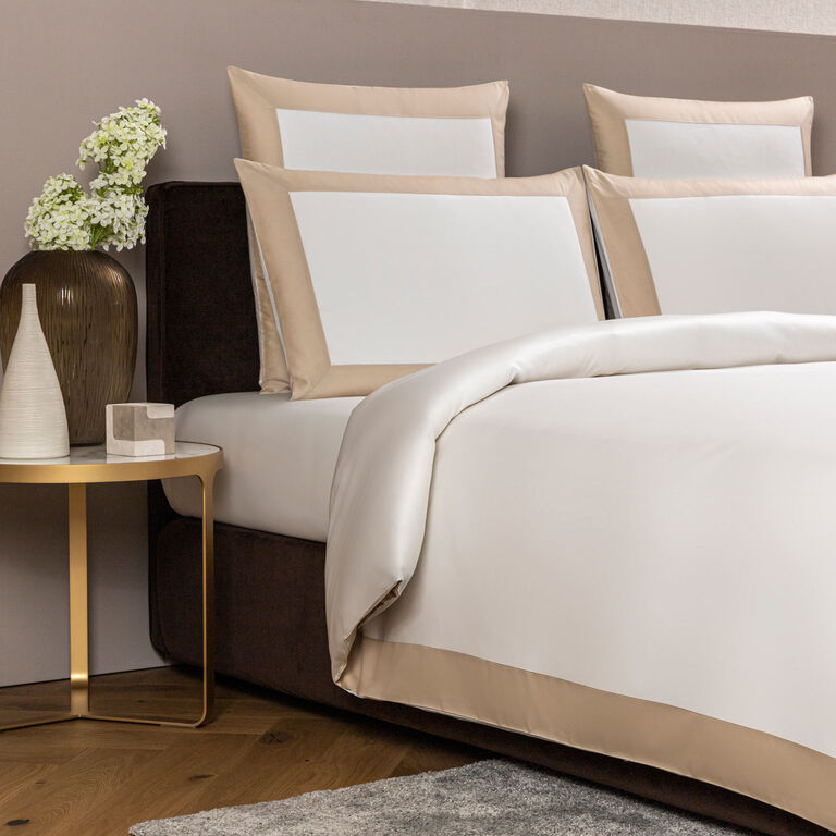 A neatly made bed features the Frette Bold | Duvet Cover in white and beige 100% cotton, four pillows, a brown upholstered headboard, and a round gold bedside table adorned with vases and flowers.
