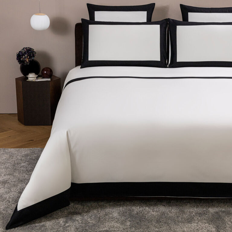 A neatly made bed showcases Frette's Bold | Duvet Cover in white with black borders, crafted from 100% cotton. Two large pillows and a matching duvet stand out, beside a table with a vase and cup. Made in Italy.