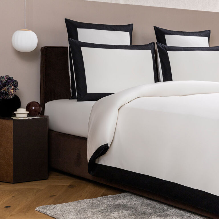 A modern bed with the Bold | Duvet Cover by Frette in 100% white cotton and black borders, styled with two large and two small pillows; nearby are a nightstand with decor and a hanging lamp.