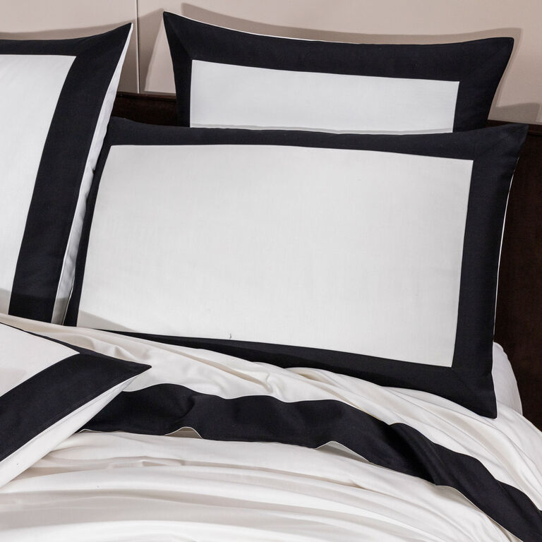 A bed is dressed with Frette’s Bold | Sleeping Sham, featuring white cotton sateen and a thick black trim for a striking pillowcase look.
