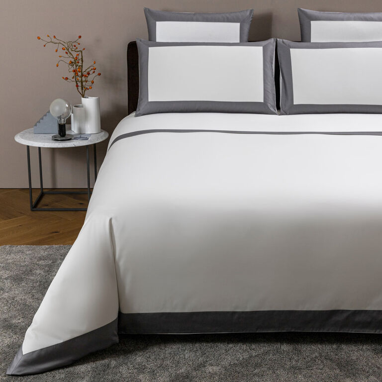 A neatly made bed features the Bold | Duvet Cover by Frette in 100% cotton, accented with white and gray bedding, matching pillows, and a bedside table displaying a lamp, a vase with branches, and a small decor piece.