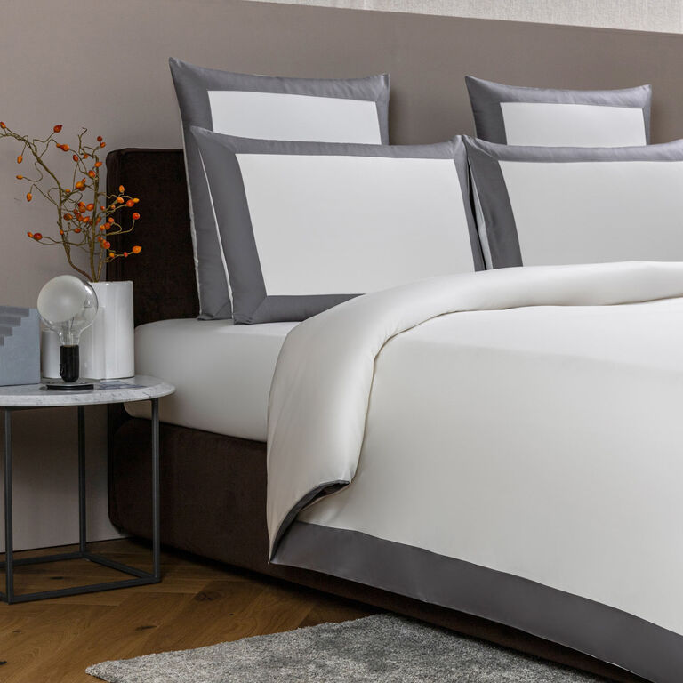 A neatly made bed features Frette’s Bold | Duvet Cover in white and gray 100% cotton bedding and pillows, next to a small round side table topped with a glass lamp, a book, and a vase of orange berries.