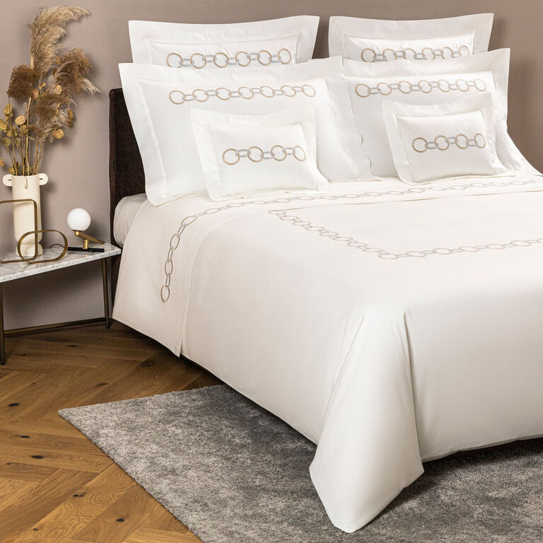 A neatly made bed features Frette's Links Embroidered Duvet Cover in white with a beige chain pattern, flanked by a marble-topped nightstand holding a lamp and decorative vase.