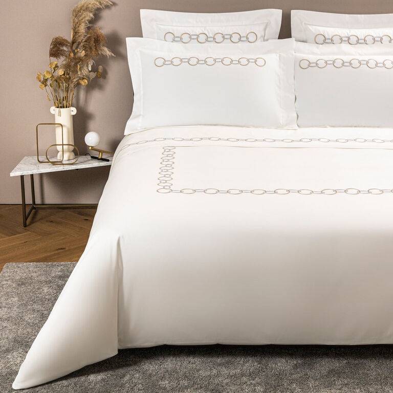 A neatly made bed features the Frette Links Embroidered Duvet Cover in white cotton sateen with a beige chain pattern, set next to a marble nightstand topped with a lamp, vase, and decorative grass.