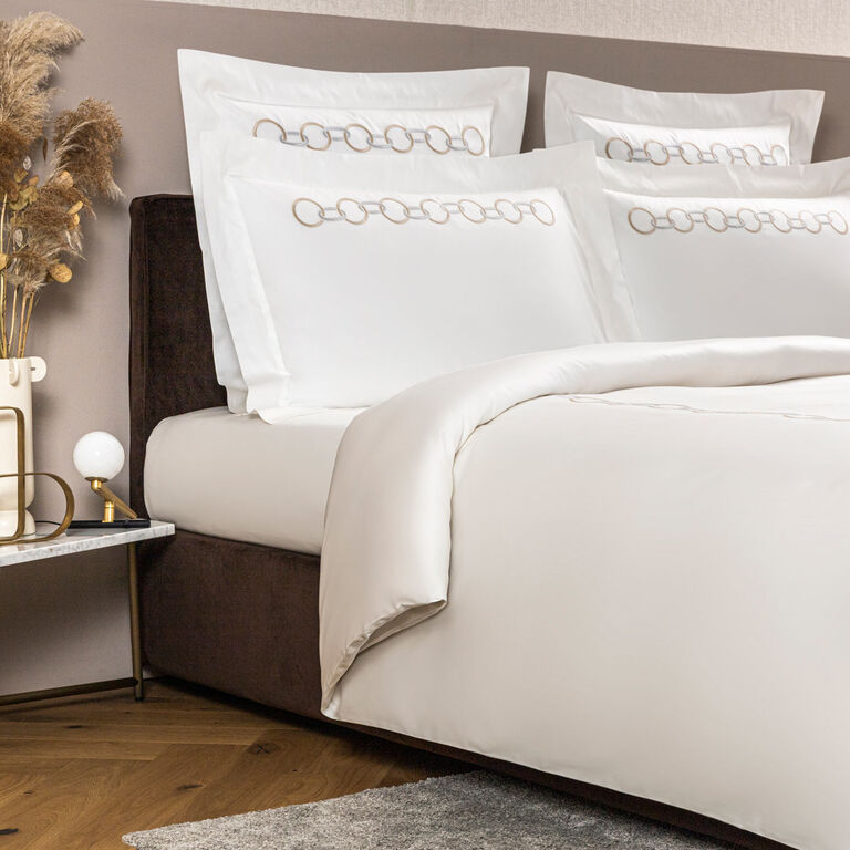 A neatly made bed features Frette’s Links Embroidered Duvet Cover and ring-patterned pillows, beside a nightstand with a lamp, vase, and dried flowers.