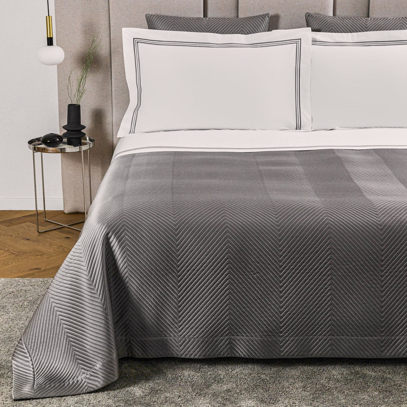 A neatly made bed features the Luxe Herringbone | Bedspread by Frette in a gray chevron pattern with two white pillows; a small round side table with decor stands nearby.