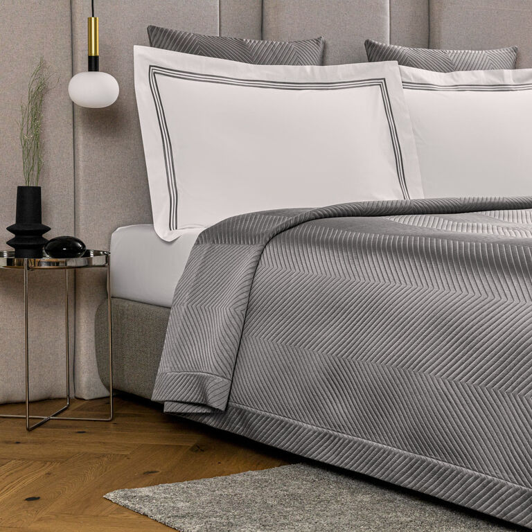 A modern bedroom features a neatly made bed with Frette’s Luxe Herringbone Bedspread and white pillows with black trim, beside a small round side table adorned with decorative items.
