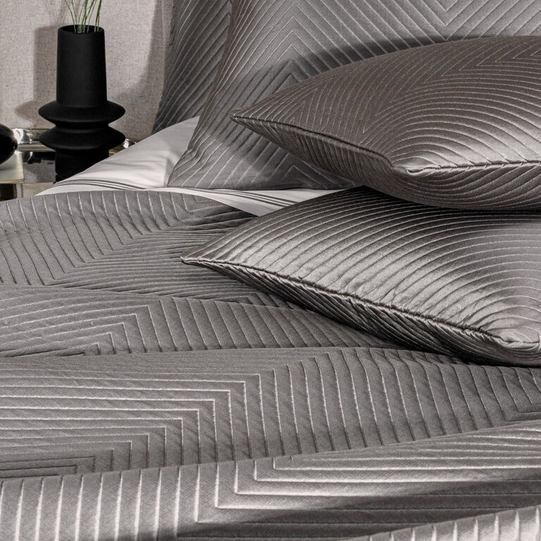 Close-up of a neatly made bed with gray, geometric pillows and a Luxe Herringbone | Decorative Sham by Frette, beside a black vase and lamp on the nightstand.