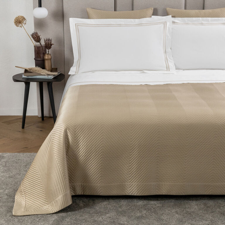 A neatly made bed with white pillows and the Frette Luxe Herringbone Bedspread in beige satin sits beside a small black side table with decor accents, placed atop a grey rug.