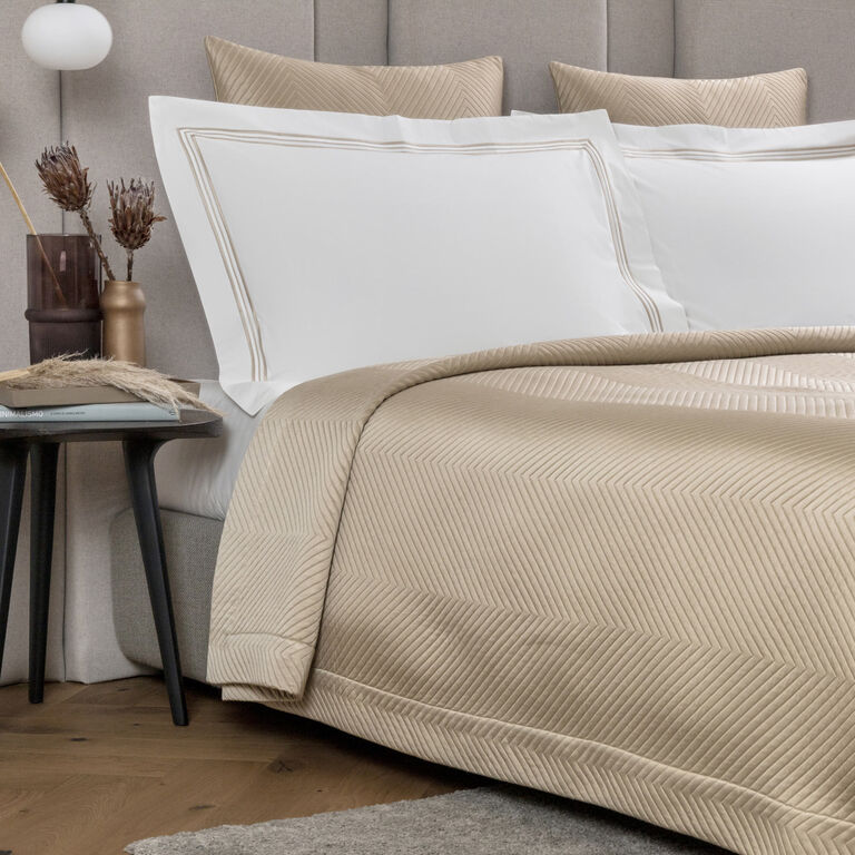 The Frette Luxe Herringbone Bedspread in beige is paired with white pillows on a neatly made bed with a gray headboard; a side table holds decorative vases and dried plants.