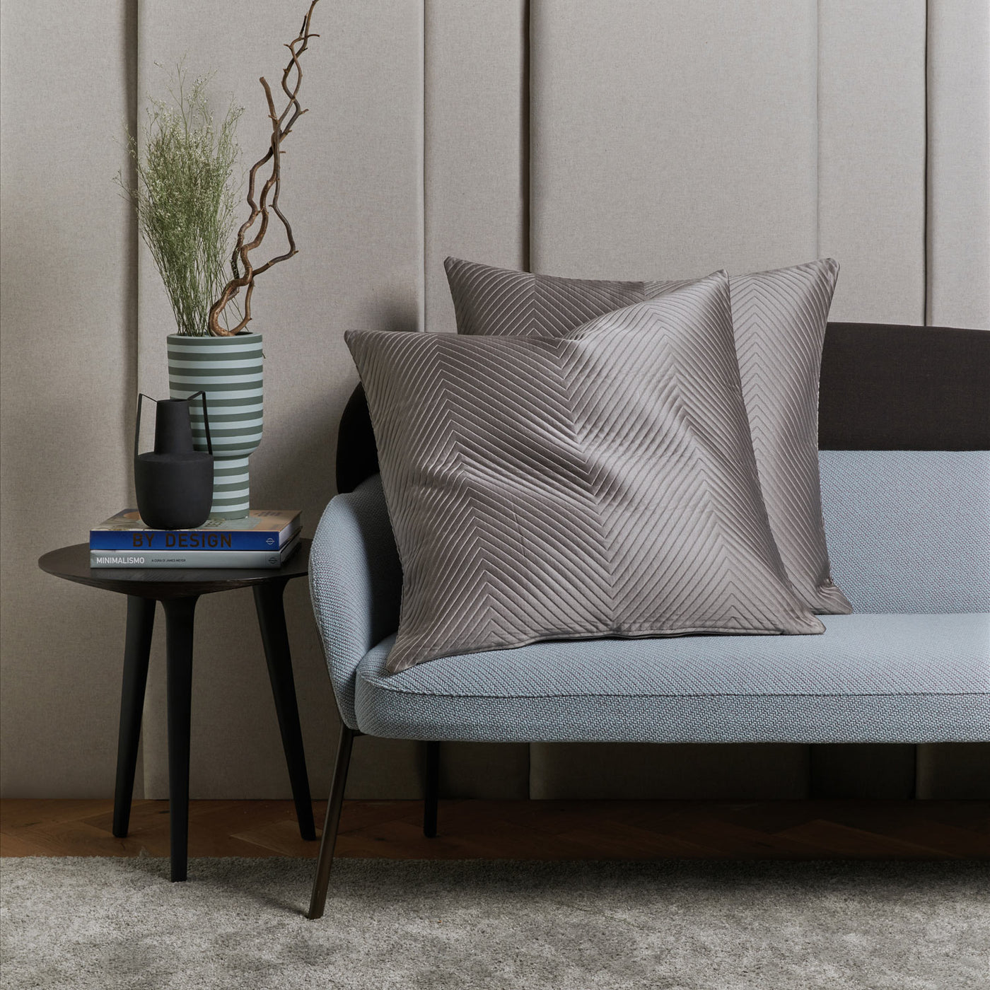 A modern living room scene with a light blue sofa, two gray textured pillows, a round black side table, a Frette Luxe Herringbone Decorative Sham draped elegantly, a vase with branches, a black mug, and a design book.