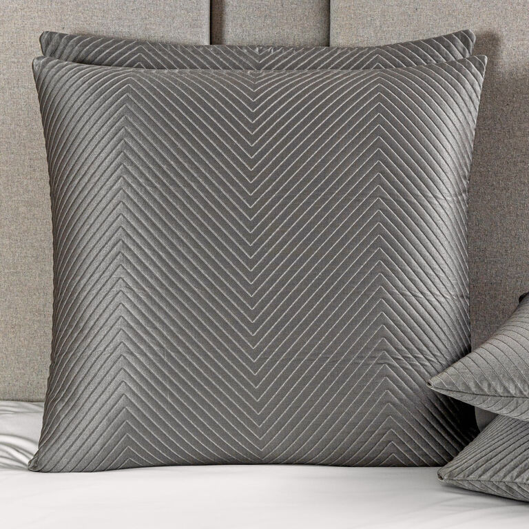 The Luxe Herringbone Decorative Sham by Frette, featuring a chevron-quilted pattern in gray, rests against a gray headboard atop a bed dressed in a luxurious silk jacquard bedcover.