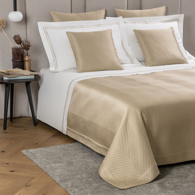 A neatly made bed with beige and white bedding, three beige square pillows, and Frette’s Luxe Herringbone Decorative Sham, next to a side table with decor on a wooden floor and gray rug.