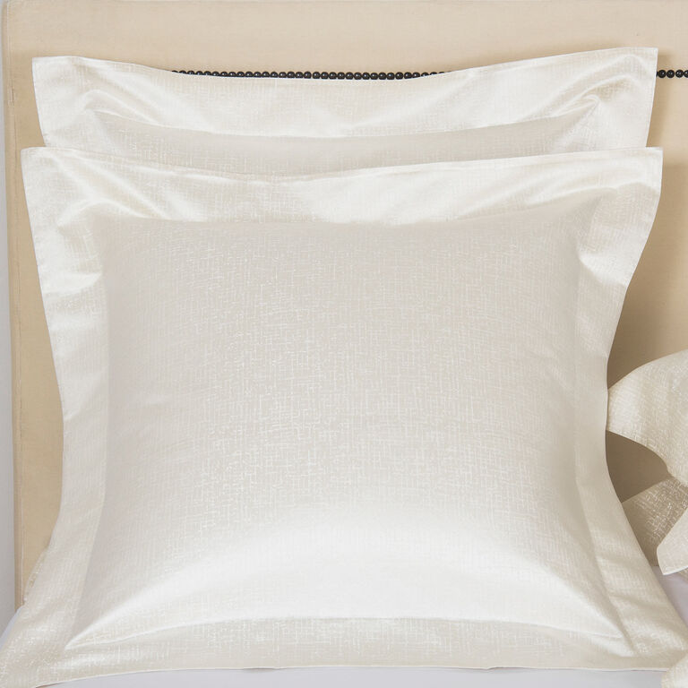 A close-up of two Luxe Glowing Weave | Shams by Frette, featuring a subtle cotton silk jacquard texture in white, set against a beige headboard.