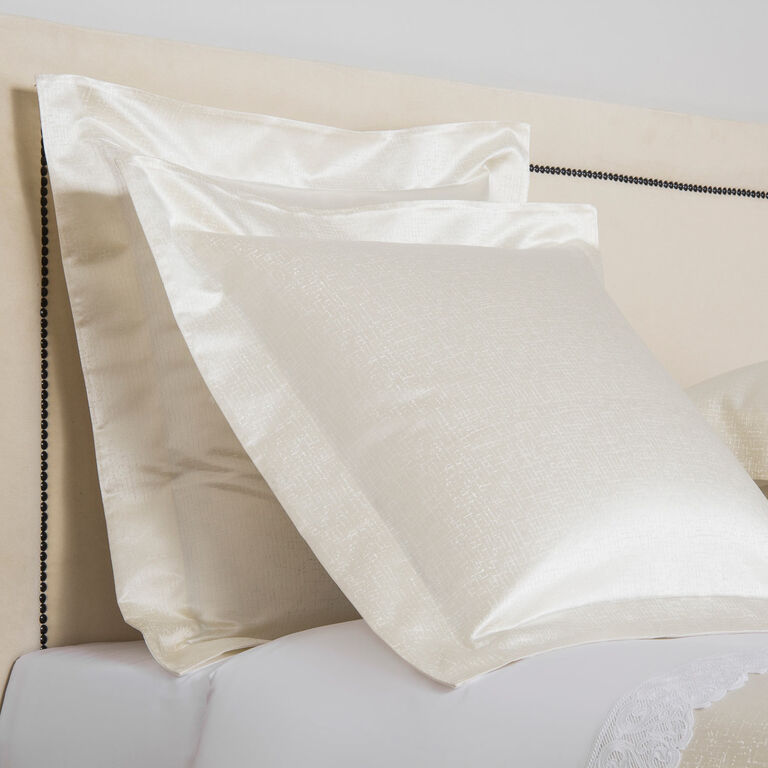 Two Luxe Glowing Weave | Shams by Frette in white cotton sateen rest against a beige upholstered, nailhead-trimmed headboard, behind crisp white bedsheets.