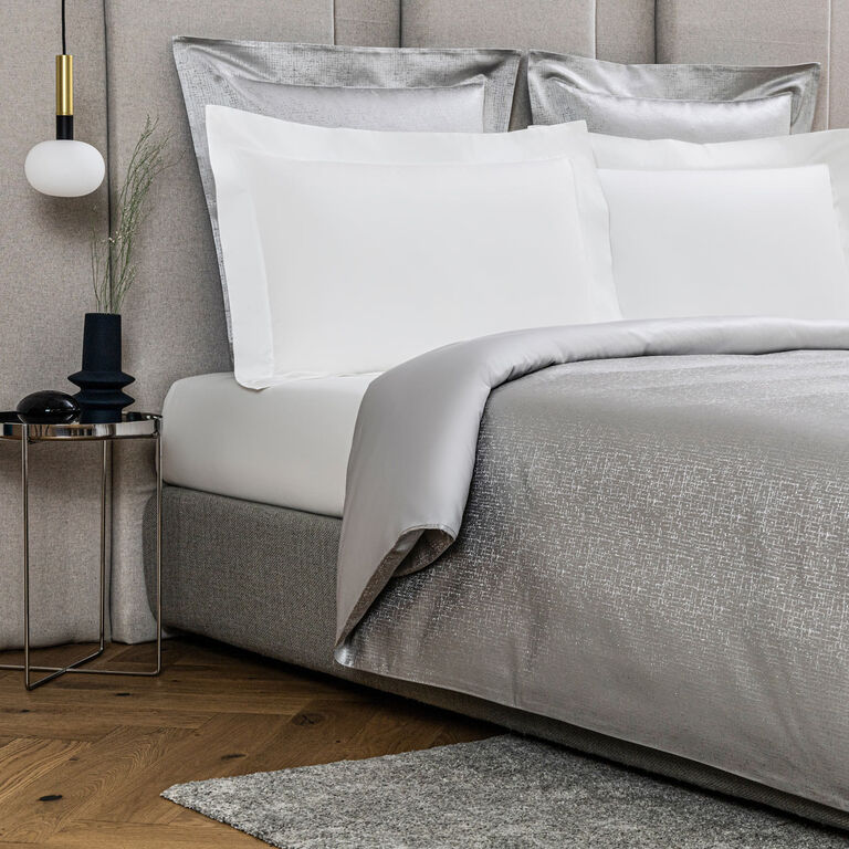 A modern bed with white pillows and the Luxe Glowing Weave Duvet Cover by Frette, beside a small round side table with a lamp, vase, and decor, on a wood floor topped with a gray rug.