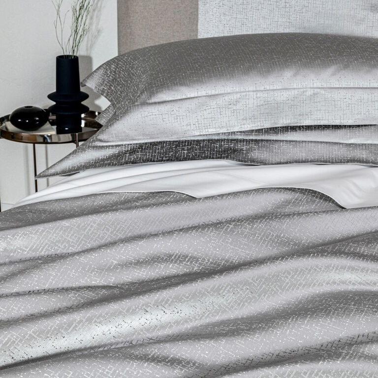 A bed dressed in Frette’s Luxe Glowing Weave shams features silver-patterned cotton silk jacquard, coordinated pillows, and crisp white sheets beside a round table with a vase, plant, and decor accents.