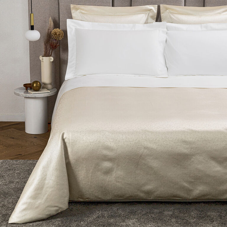 A neatly made bed with white pillows and sheets, topped with the Frette Luxe Glowing Weave Duvet Cover in a beige satin finish. A bedside table with decor sits next to the bed.