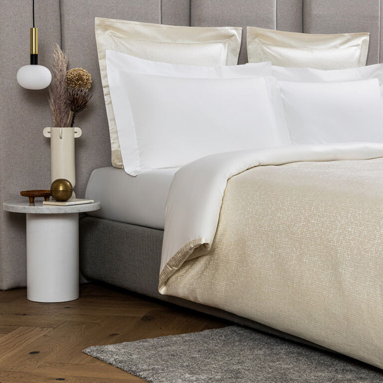 A bed styled with the Frette Luxe Glowing Weave Duvet Cover stands next to a round side table holding a vase of dried plants, a lamp, and decor, set on a wooden floor.