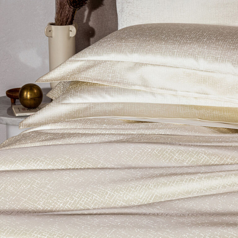 A neatly made bed displays Frette’s Luxe Glowing Weave shams—cream-colored cotton sateen with a subtle geometric pattern—beside a white nightstand adorned with decorative accents.