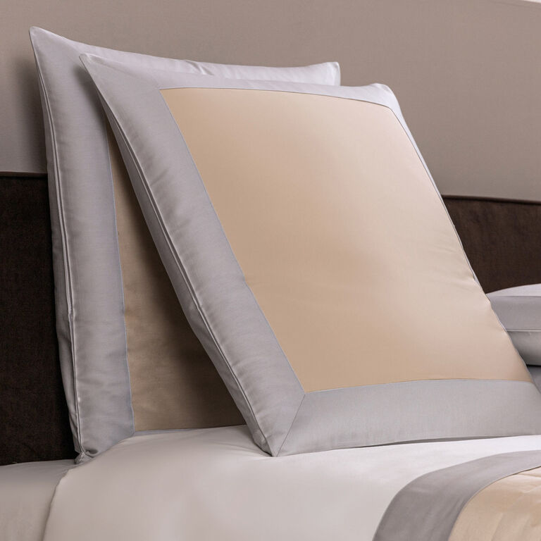 Two rectangular Frette Euro Shams with beige centers and light gray borders rest upright against a headboard on a neatly made bed, complemented by a lightweight quilt and a luxurious sham for added comfort and style.