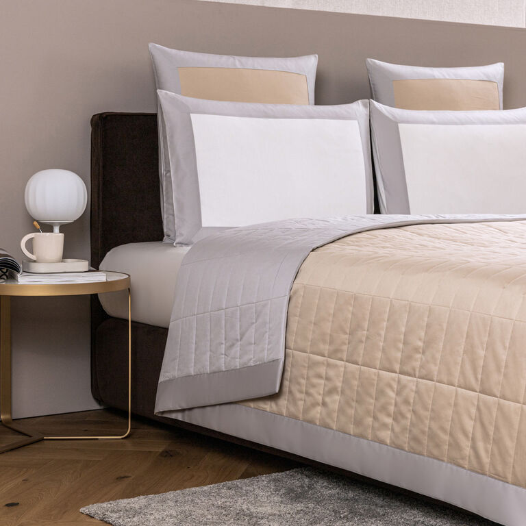 A modern bed with beige and gray bedding, accented pillows, and the Frette Rectangular Light Quilt in cotton sateen, complemented by a round side table holding a cup, saucer, book, and small lamp on a wooden floor.