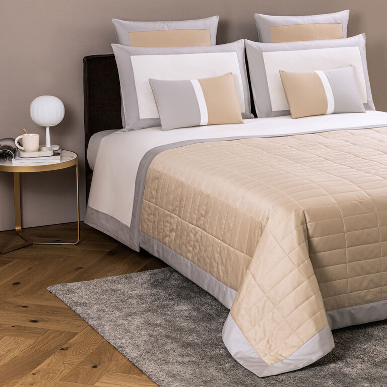 A neatly made bed with beige and gray bedding, multiple pillows, and a Frette Rectangular Light Quilt in soft cotton sateen, beside a round side table with a lamp, cup, and saucer on a wooden floor and gray rug.