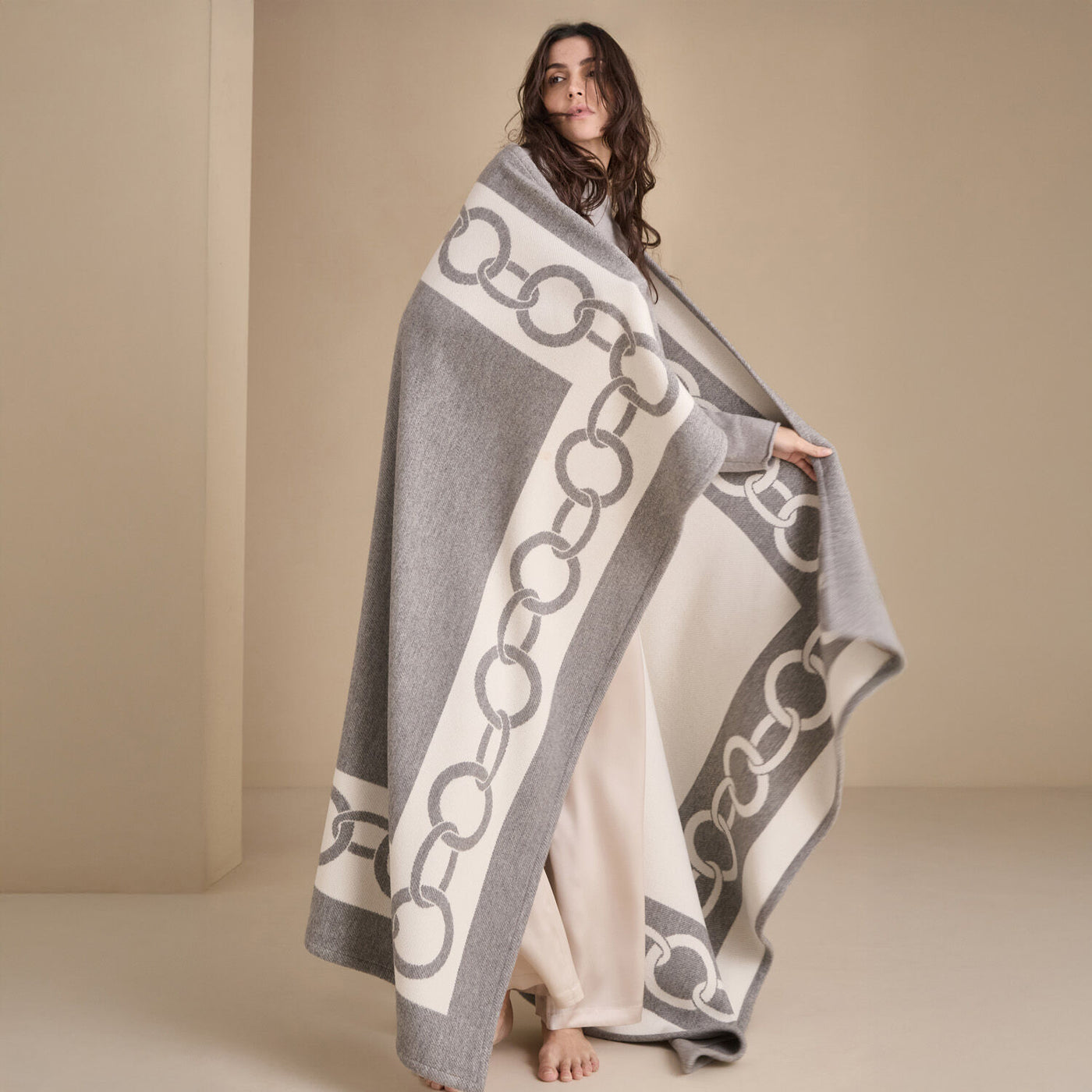 A barefoot woman is draped in Frette's The Chains Throw Blanket, featuring a gray and white chainlink pattern, set against a neutral background.