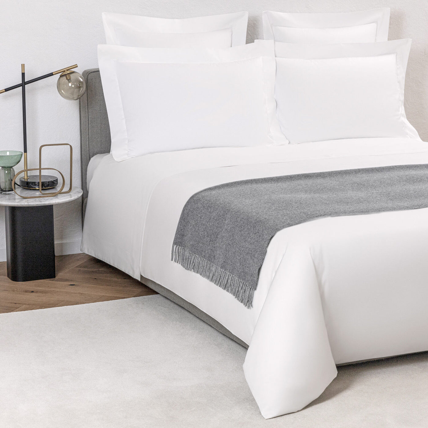 A neatly made bed with white bedding and pillows is accented by The Pure Cashmere Throw from Frette at the foot, alongside a black nightstand with a lamp and decor.