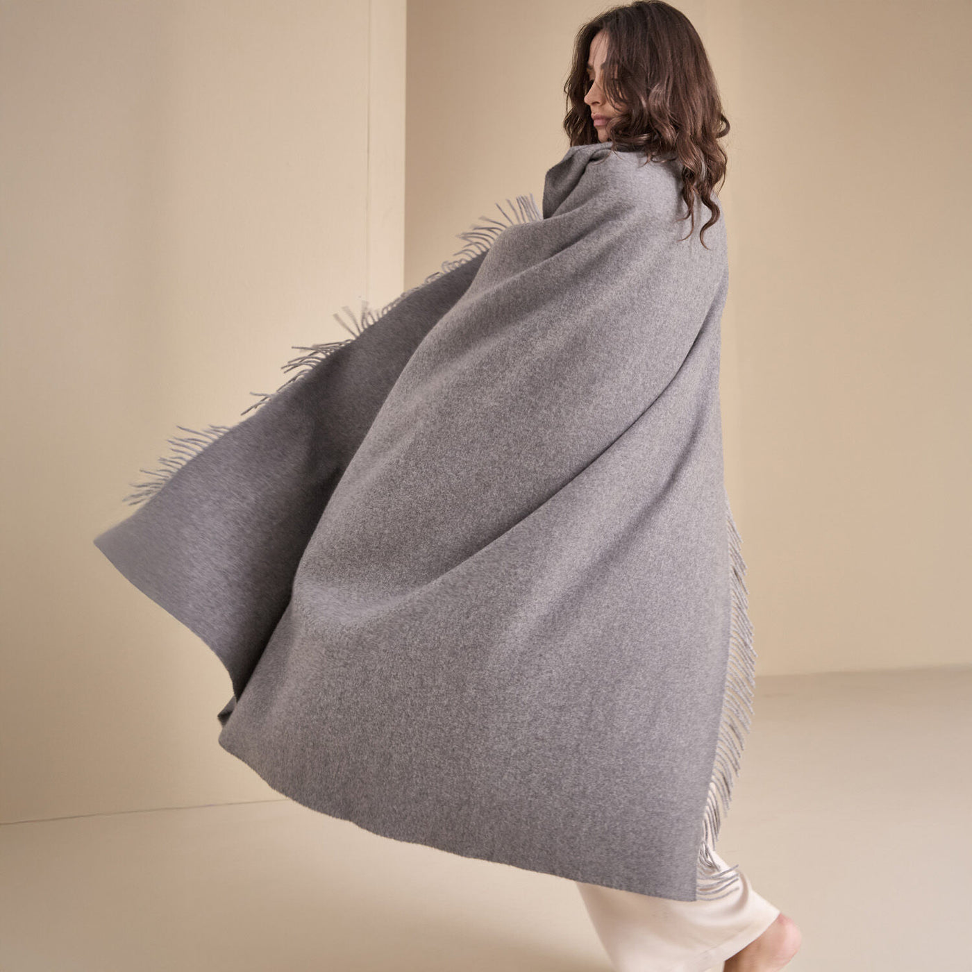 A person stands barefoot on a light-colored floor, wrapped in Frette’s The Pure Cashmere Throw in large grey, made in Italy, against a neutral background.