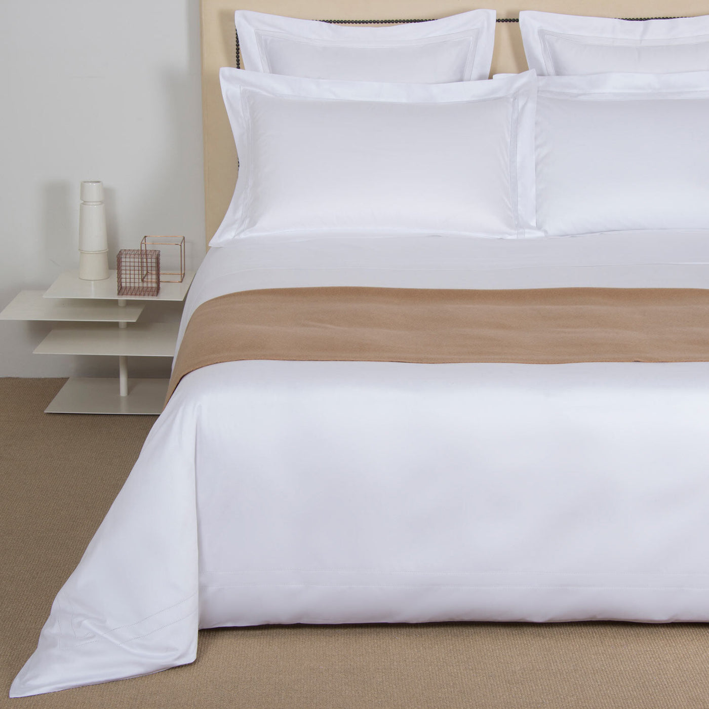 Made bed with white sheets, four pillows, and The Pure Cashmere Throw by Frette, beside a small white table with a carafe and wire basket.