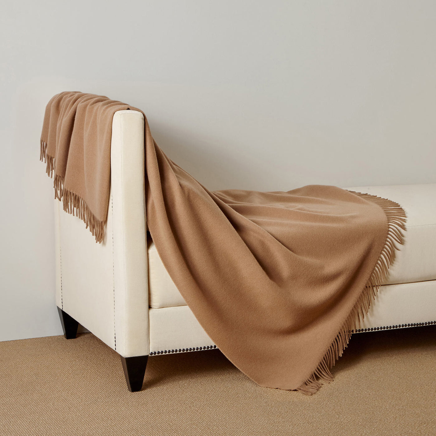 The Pure Cashmere Throw by Frette, featuring fringed edges, is draped over a cream-upholstered bench with black wooden legs in a neutral room.