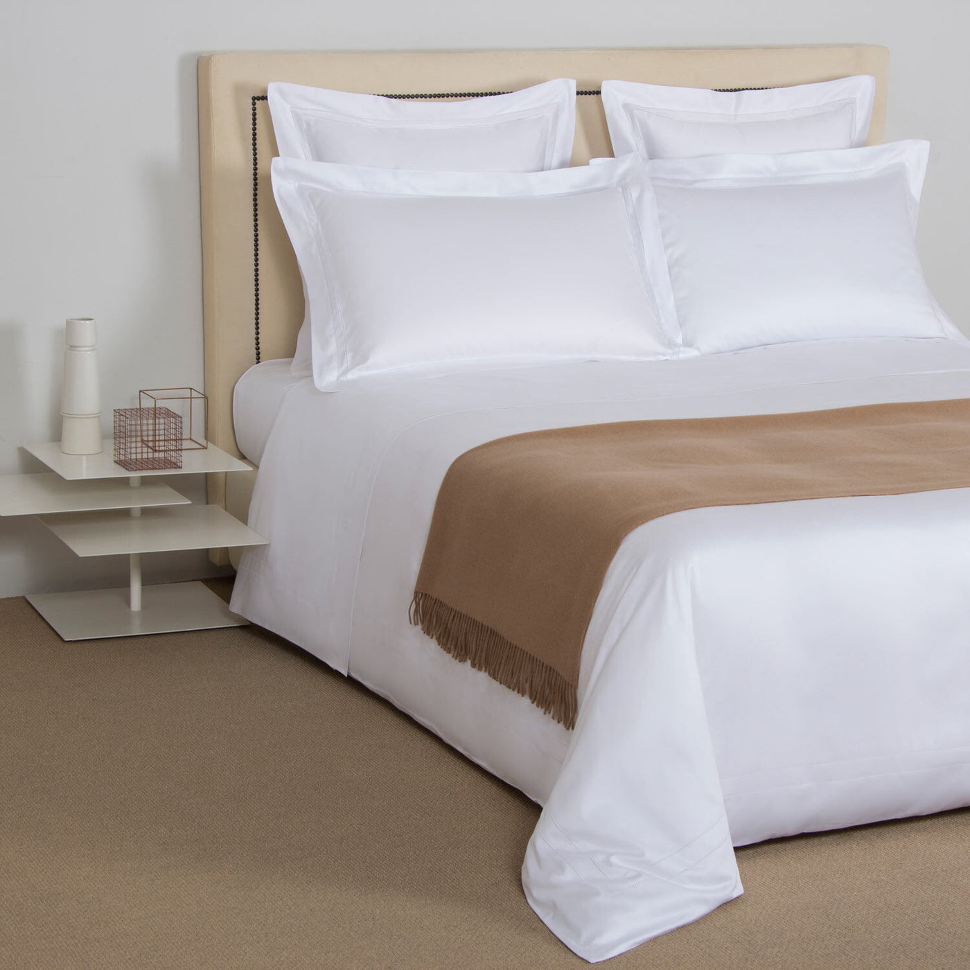 A neatly made bed with white sheets, four white pillows, and The Pure Cashmere Throw by Frette at the foot, next to a small modern white nightstand adorned with decorative items.