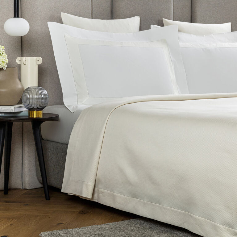 A bed dressed in Frette's Cavalry | Bedspread in white cotton jacquard, topped with pillows, sits next to a dark bedside table featuring a beige vase with flowers, a lamp, and books.