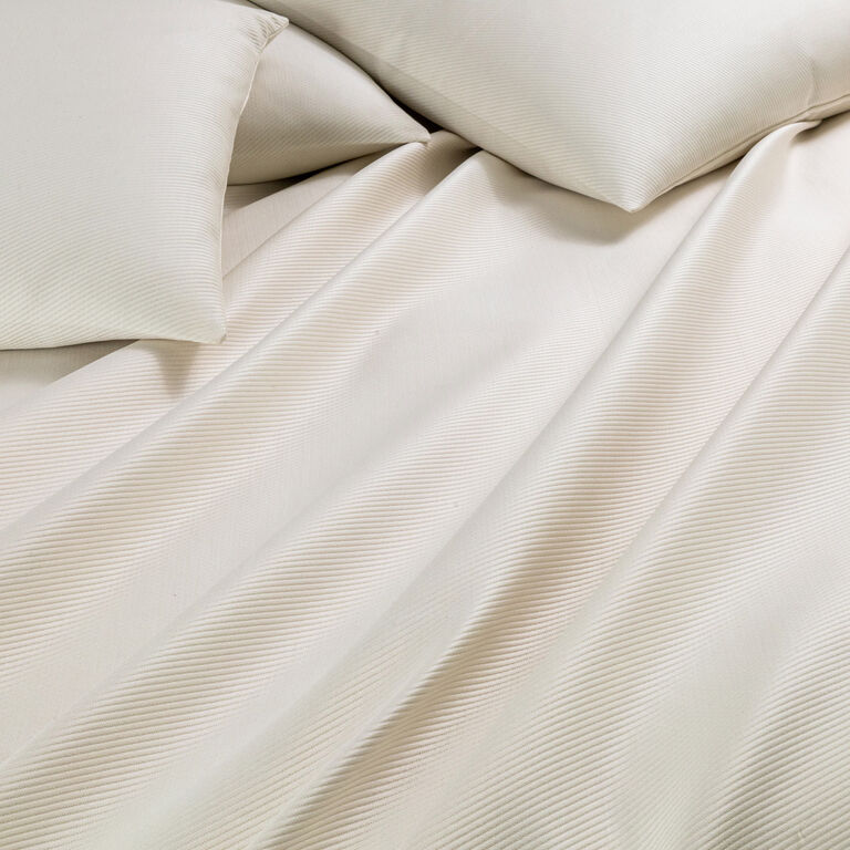 Close-up of an unmade bed featuring the Cavalry Bedspread by Frette, in cream-colored textured cotton jacquard with two matching pillows. Soft folds and subtle diagonal lines add elegance to the luxurious bedding.
