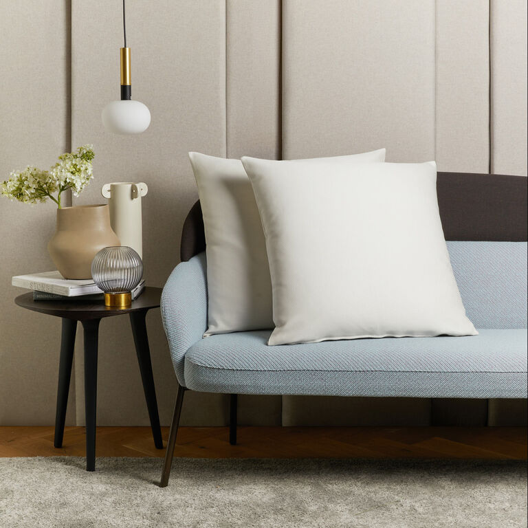 A modern living room with a blue and gray sofa, two white Frette Cavalry Euro Shams, a black side table with a vase of flowers, a decorative object, books, and a hanging pendant light blends classic style with contemporary comfort.