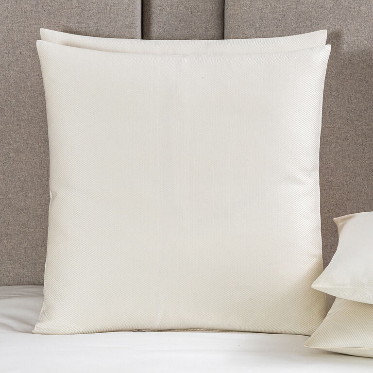 A large cream-colored Cavalry | Euro Sham by Frette is propped upright on a bed with a beige upholstered headboard, next to a matching pillow, creating a classic accent reminiscent of the Cavalry collection.