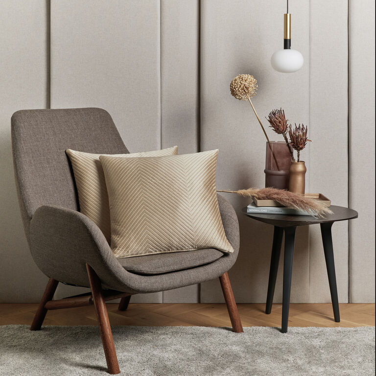 Modern armchair with two beige patterned cushions beside a black side table holding vases, dried flowers, books, and a hanging white pendant light—add sophistication with Frette's Luxe Herringbone Decorative Sham.