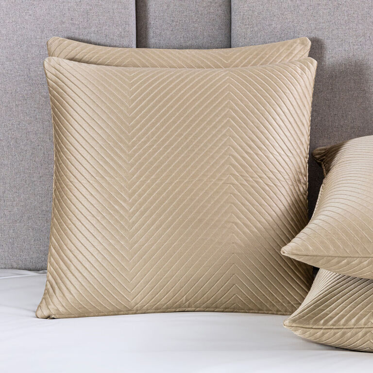 Two beige, quilted, square pillows with a chevron pattern rest on a bed topped with the Frette Luxe Herringbone Decorative Sham and a light gray upholstered headboard. A third pillow appears partially visible on the right.