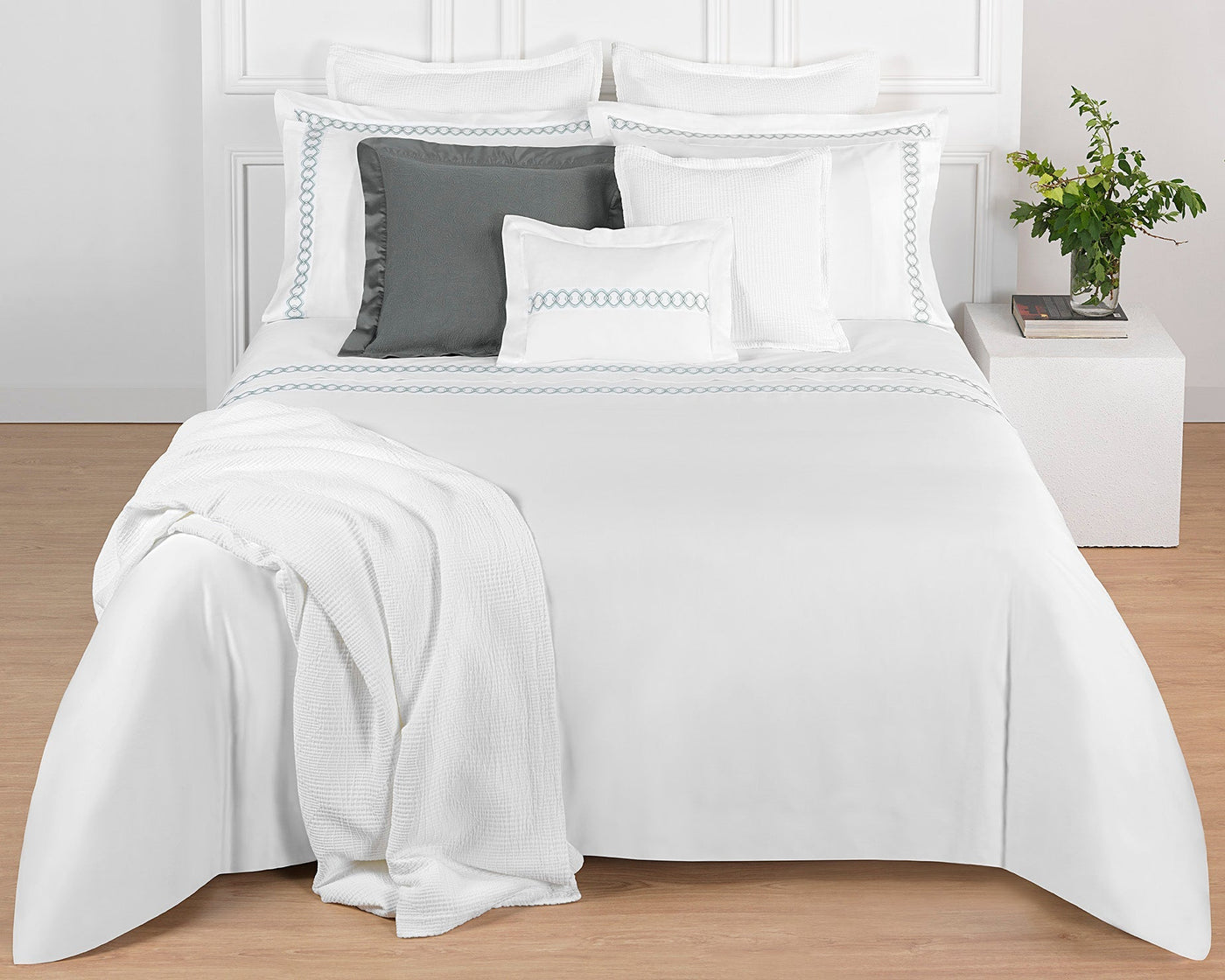 A neatly made bed with white luxury bedding, accented by Amalia Home Collection Ornato Boudoir pillows in white and gray, a folded Egyptian Cotton blanket at the foot, and a small side table with a glass vase of greenery.
