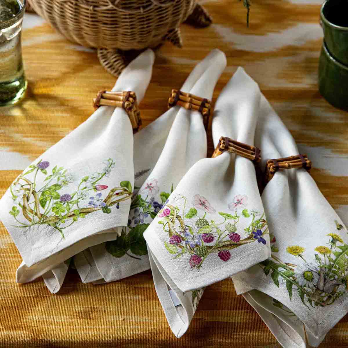 Four Juliska Meadow Walk napkins with varied botanical prints, rolled in bamboo rings on a striped table.
