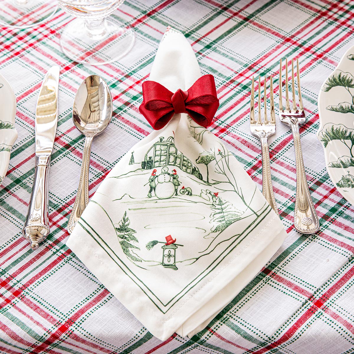 Evergreen Winter Frolic napkin with a red bow at a place setting on a plaid tablecloth
