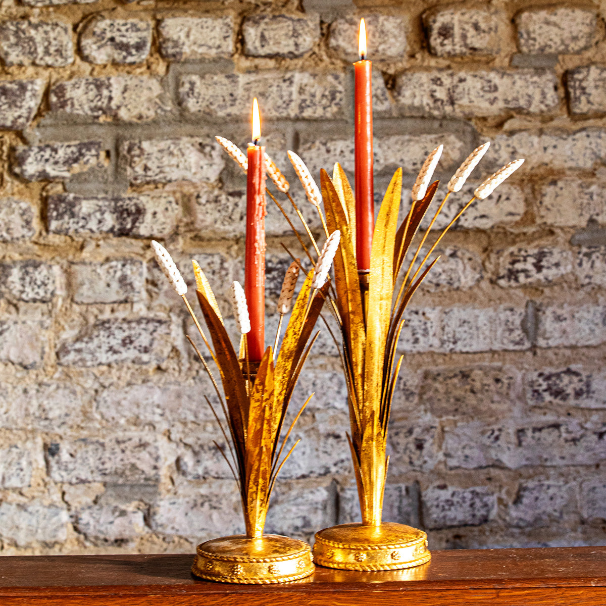 Two Forest Walk gold wheat candlestick holders with red tapers against a brick wall