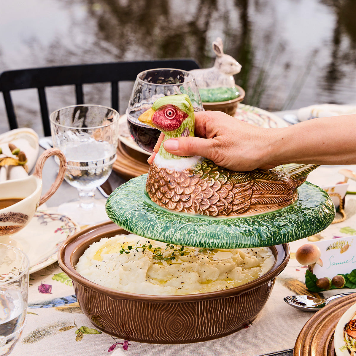 Hand lifting the pheasant lid off a Forest Walk baker of mashed potatoes on an outdoor table