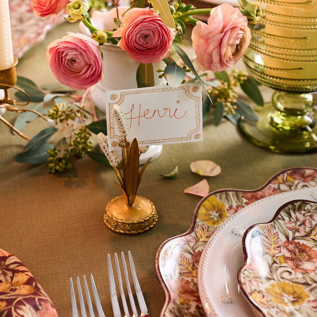Forest Walk gold wheat place card holder with a card reading Henri beside pink ranunculus